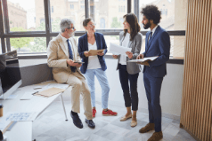 3 people stand for an office meeting whilst one sits on a desk
